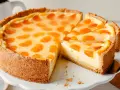 Ein saftiger Schmandkuchen mit Mandarinen, der auf einem weißen Tortenständer präsentiert wird. Ein Stück wurde bereits entnommen.