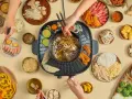 Hände greifen nach Zutaten für einen Hot Pot. Fleisch, Gemüse, Garnelen und Nudeln werden in die köchelnde Brühe getaucht.