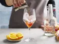 Zabawne przygotowanie Aperol Spritz Rezept Original. Likier wlewa się do kieliszka z lodem. Obok leżą pomarańcze i butelka prosecco.