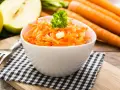 Ein frischer Karotten-Apfel-Salat, garniert mit Petersilie, steht auf einem karierten Tuch. Im Hintergrund liegen ganze Karotten und ein halber Apfel.
