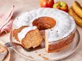 Saftiger Apfel-Bananen-Kuchen, bestäubt mit Puderzucker. Daneben liegen frische Äpfel und Bananen.