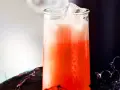 Erfrischender Aperol Sour mit Eiswürfeln in einem Glas, daneben ein dunkler Becher und Spinnweben-Deko.
