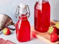 Zwei Flaschen mit leuchtend rotem Erdbeersirup, frisch zubereitet. Frische Erdbeeren liegen daneben.