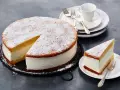Eine köstliche Quark Sahne Torte, mit Puderzucker bestäubt, wartet auf ihre Genießer. Ein Stück ist bereits auf einem Teller angerichtet.