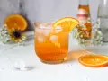 Erfrischender Crodino Spritz mit Eiswürfeln und Orangenscheibe, dekoriert mit kleinen weißen Blüten.