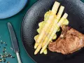 Ein saftiges Schweinekotelett liegt neben einem Haufen cremigen Spargel in der Pfanne, beträufelt mit einer würzigen Sauce.