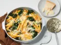 Herzhafter nudelauflauf mit spinat, überbacken mit Käse. Daneben ein Stück Parmesan und ein dekoratives Servierbesteck.