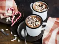 Zwei Tassen heißer Kakao mit Marshmallows und Schokostückchen, daneben Zuckerstangen und eine karierte Serviette.