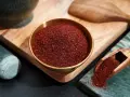 Schüssel mit gemahlenem Sumak, daneben eine Holzschaufel mit dem Gewürz. Mörser und Stößel im Hintergrund.