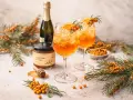 Zwei leuchtend orangefarbene Sanddorn Spritz-Cocktails mit Eis, garniert mit Orangenscheiben und Sanddornzweigen. Daneben ein Glas Sanddorn & Honig.