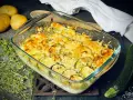 Goldbrauner Zucchini-Kartoffel-Auflauf mit Käse überbacken, garniert mit frischem Thymian. Daneben liegen Kartoffeln und eine Zucchini.