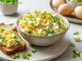 Cremiger Eiersalat mit Joghurt, garniert mit Frühlingszwiebeln, auf einer Scheibe Toastbrot. Daneben eine Schüssel voller Salat und einige gekochte Eier.