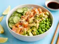 Schüssel mit einem frischen Salat aus Lachs, Avocado, Edamame und Gurkenscheiben, beträufelt mit cremiger Sauce und Sesam. Zitronenspalten und Sojasauce daneben.