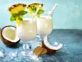 Zwei cremige Malibu Cocktails mit Ananas und Minze, umgeben von Kokosnuss und Eis.