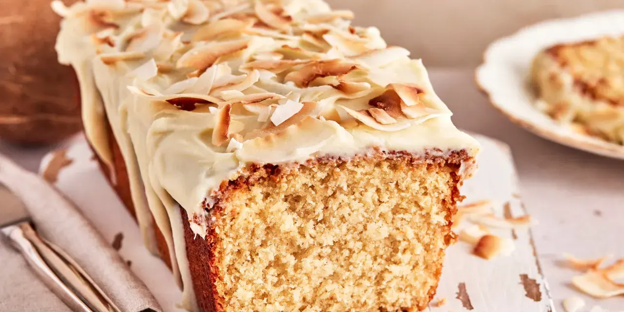 Saftiger Kuchen mit Kokosmilch, überzogen mit weißer Glasur und garniert mit gerösteten Kokoschips. Daneben eine Kokosnuss.