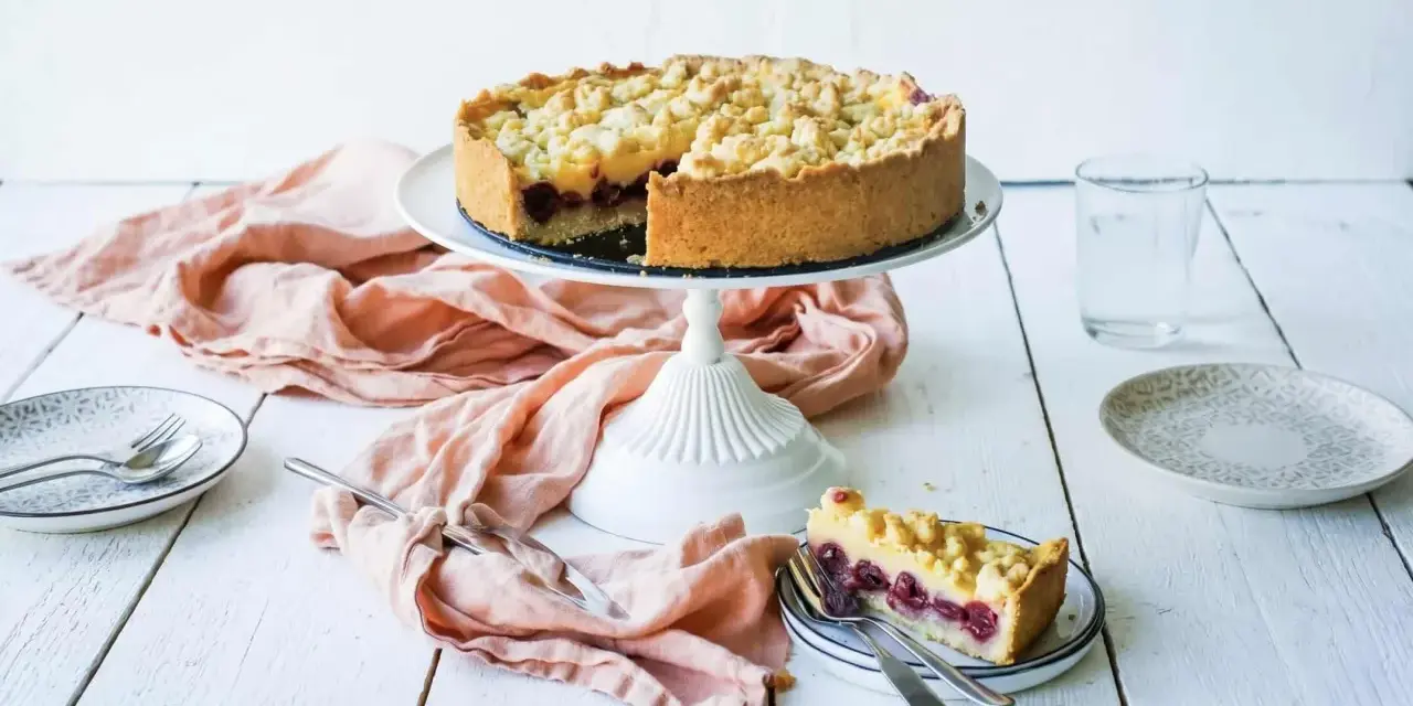 Ein saftiger Kirschkuchen mit Pudding und Streuseln auf einem Tortenständer. Ein Stück ist bereits auf einem Teller angerichtet.