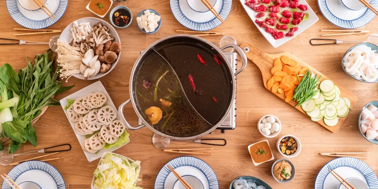 Zutaten für ein köstliches Hot Pot selber machen: Fleisch, Pilze, Gemüse, Garnelen und verschiedene Dips auf einem Holztisch arrangiert.