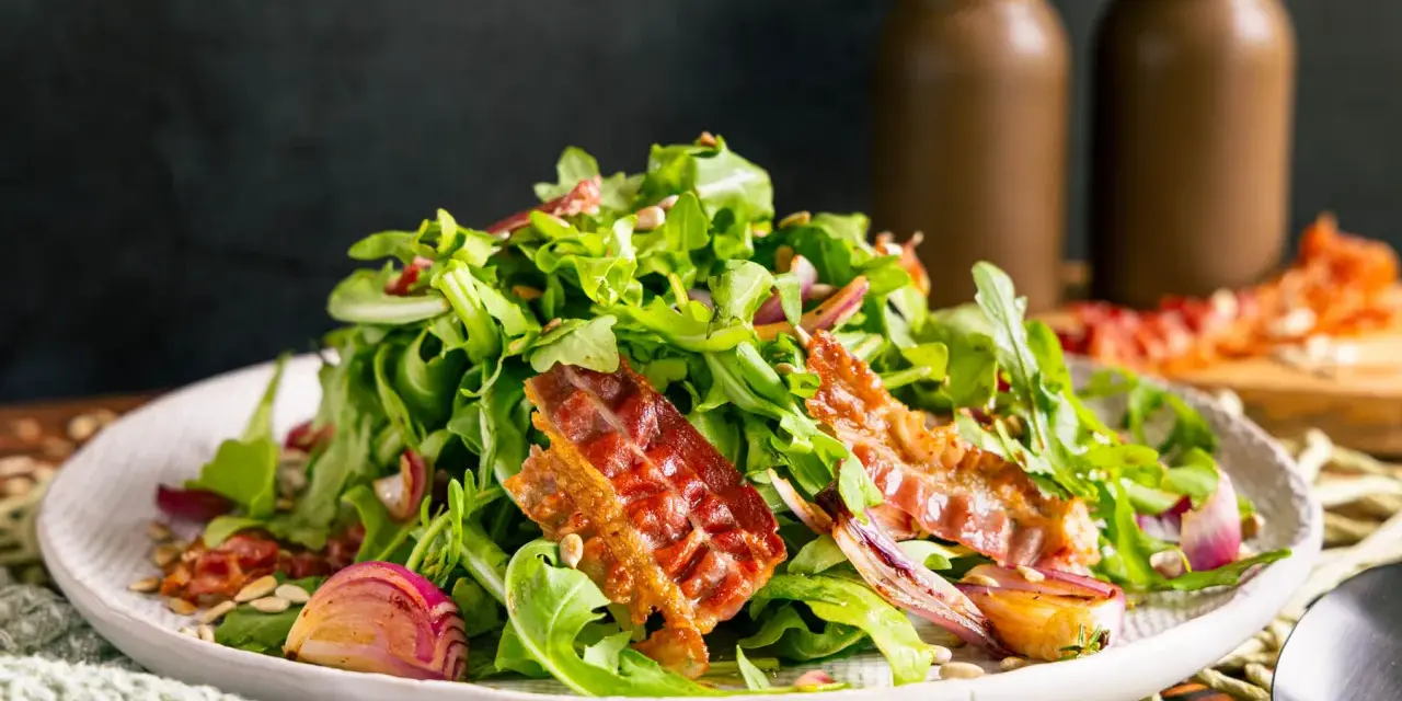 Warmer Rucolasalat mit knusprigem Speck, gerösteten Zwiebeln und Sonnenblumenkernen. Ein köstliches Gericht für Rucola Rezepte warm.
