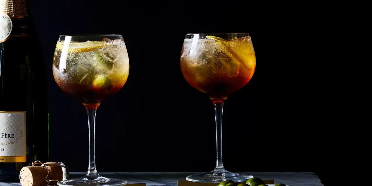 Zwei erfrischende Cynar Spritz Cocktails mit Eis, Zitrone und Oliven, daneben eine Champagnerflasche.