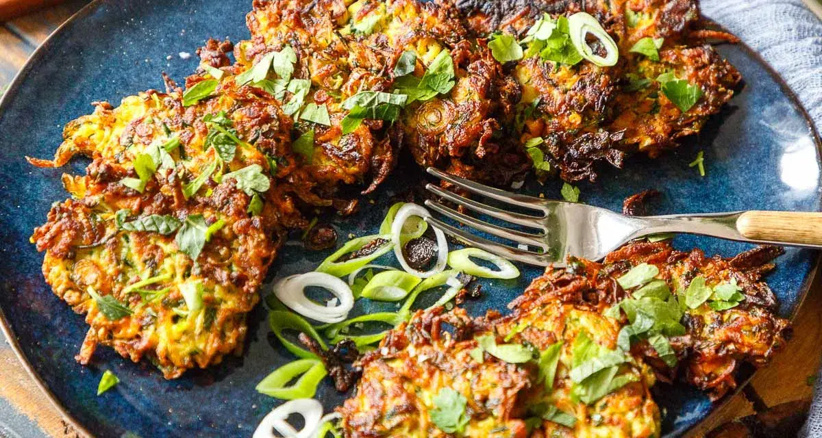 Knusprige Zucchini Kartoffel Puffer, garniert mit frischen Kräutern und Frühlingszwiebeln, liegen auf einem blauen Teller. Eine Gabel liegt daneben.