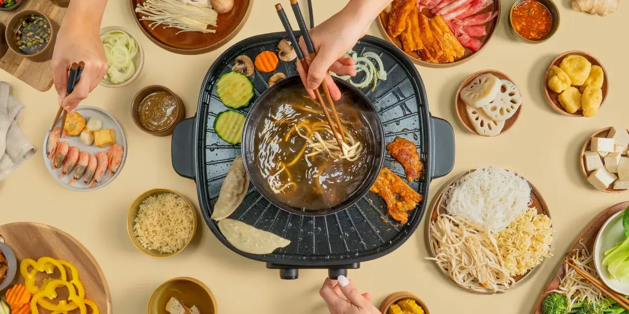 Hände greifen nach Zutaten für einen Hot Pot. Fleisch, Gemüse, Garnelen und Nudeln werden in die köchelnde Brühe getaucht.