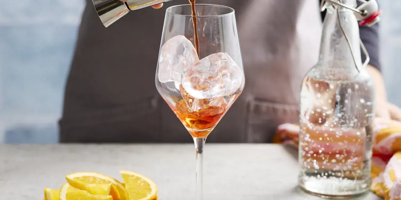 Zabawne przygotowanie Aperol Spritz Rezept Original. Likier wlewa się do kieliszka z lodem. Obok leżą pomarańcze i butelka prosecco.