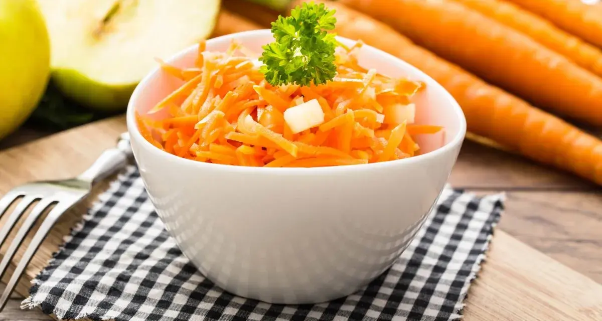 Ein frischer Karotten-Apfel-Salat, garniert mit Petersilie, steht auf einem karierten Tuch. Im Hintergrund liegen ganze Karotten und ein halber Apfel.