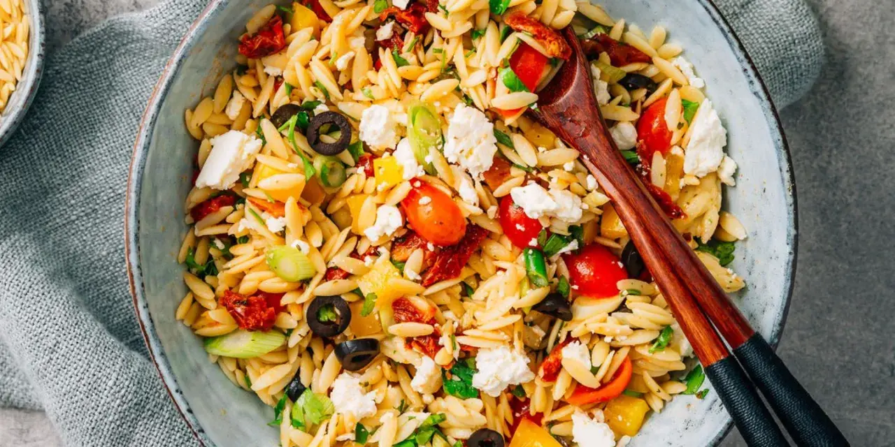 Bunter Kritharaki Salat mit Tomaten, Oliven, Feta und Kräutern, serviert in einer Schüssel mit Holzstäbchen.