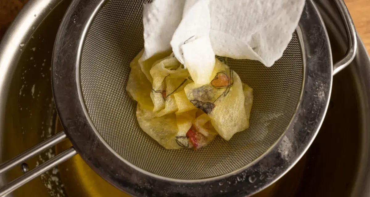Ein Sieb hält ein Stück Papier mit Blumenmuster, das über einem Topf mit geschmolzener Butter liegt. Perfekt für raffinierte Butter Rezepte.