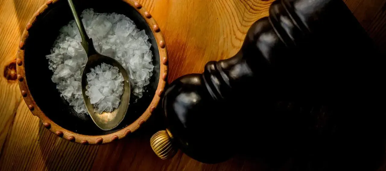 Fleur de sel, zarten Salzkristallen auf einem Löffel in einer Schale, neben einer dunklen Pfeffermühle.