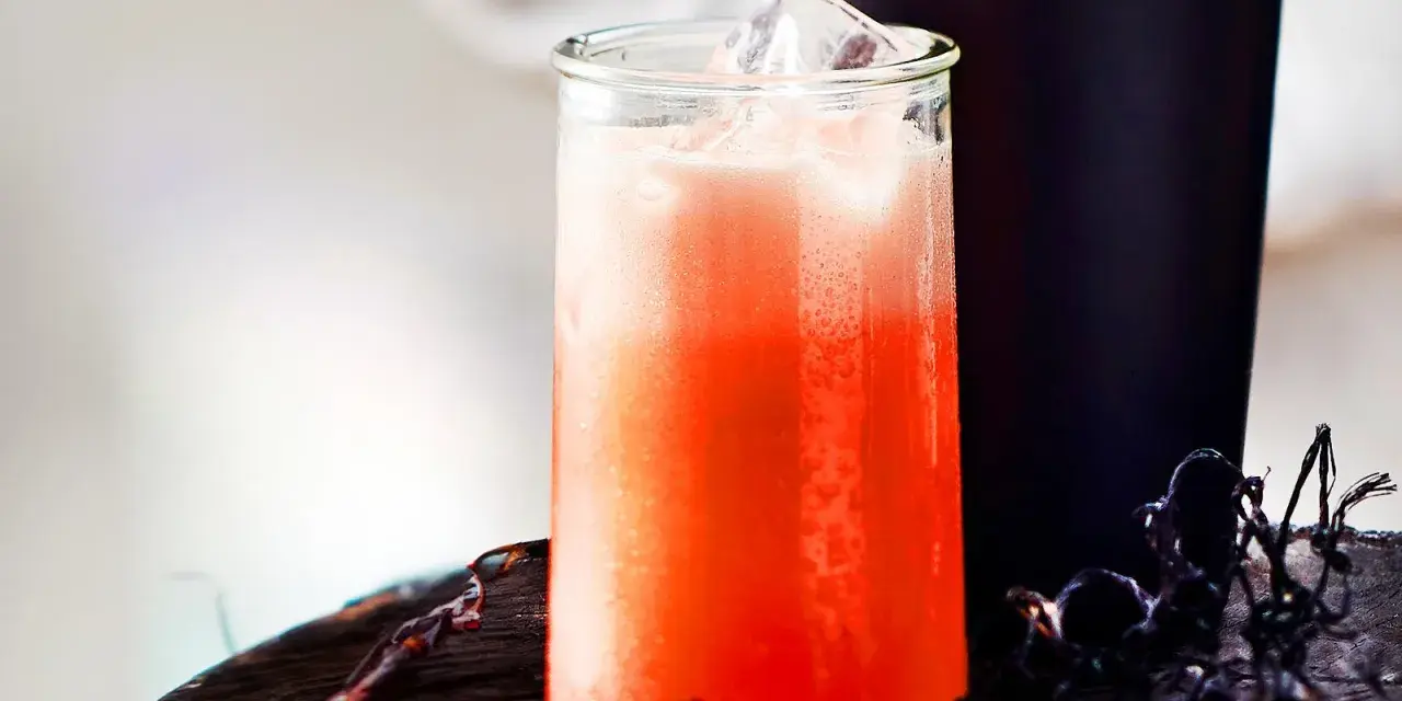 Erfrischender Aperol Sour mit Eiswürfeln in einem Glas, daneben ein dunkler Becher und Spinnweben-Deko.