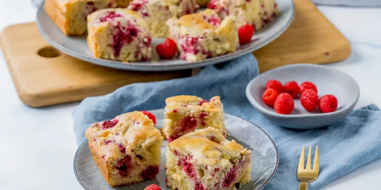Saftiger Kuchen mit Himbeeren, auf Tellern und einer Platte angerichtet, daneben eine Schale mit frischen Himbeeren.