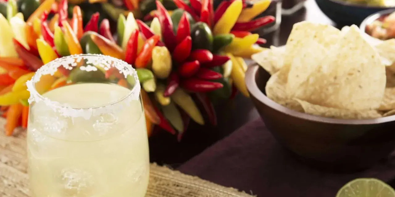 Ein erfrischendes Margarita-Rezept: Ein Glas mit Salzrand, Eiswürfeln und Limette, daneben bunte Chilis, Guacamole und Tortilla-Chips.