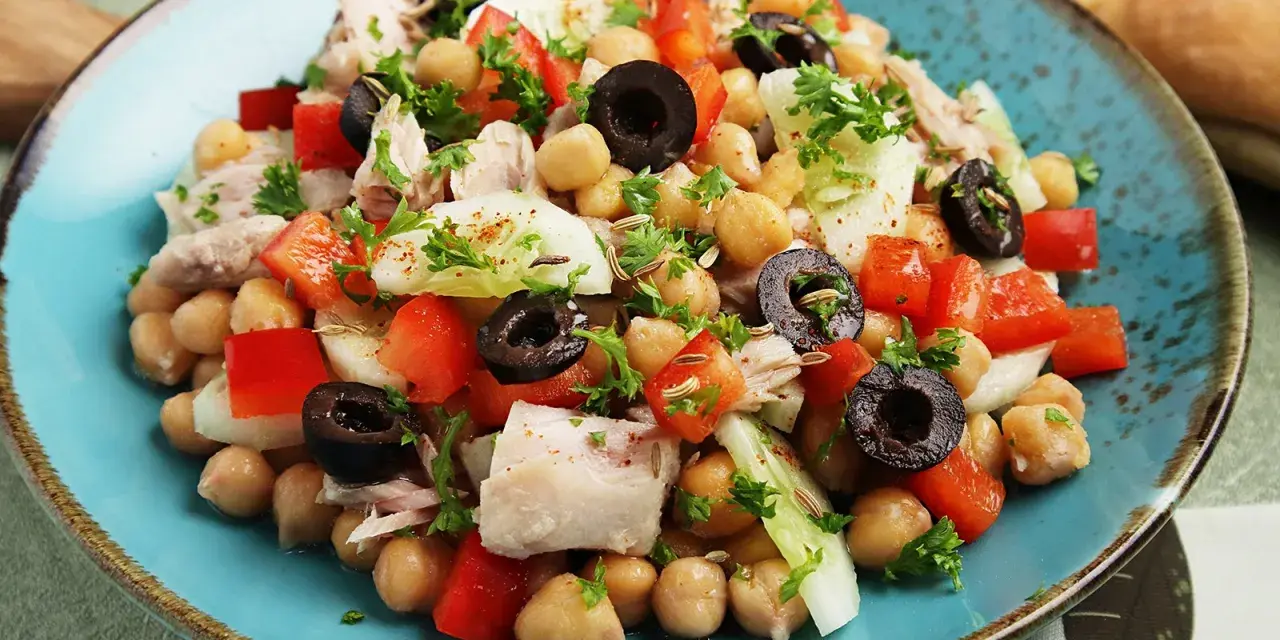Ein bunter Kichererbsen Thunfisch Salat mit Paprika, Oliven und Petersilie, appetitlich auf einem blauen Teller angerichtet.