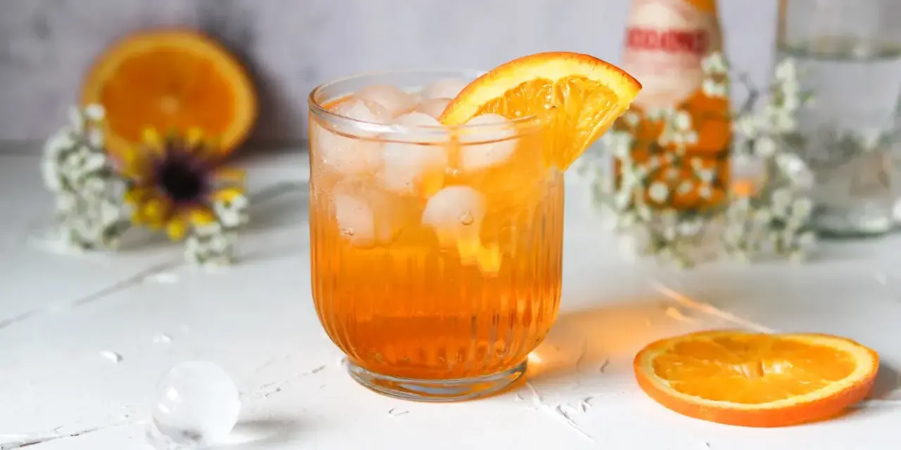 Erfrischender Crodino Spritz mit Eiswürfeln und Orangenscheibe, dekoriert mit kleinen weißen Blüten.