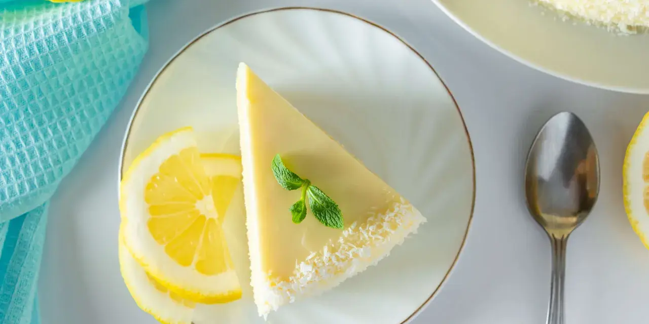 Ein Stück Philadelphia Torte Zitrone, garniert mit Minze und Kokosraspeln, neben Zitronenscheiben und einem Löffel.