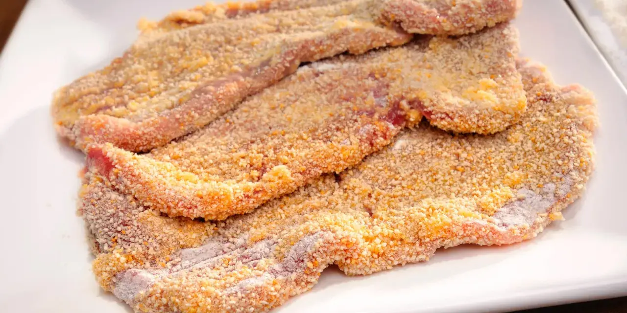 Mehrere rohe Schnitzel liegen auf einem weißen Teller, bereit zum Schnitzel panieren. Die Panade ist goldbraun und knusprig.