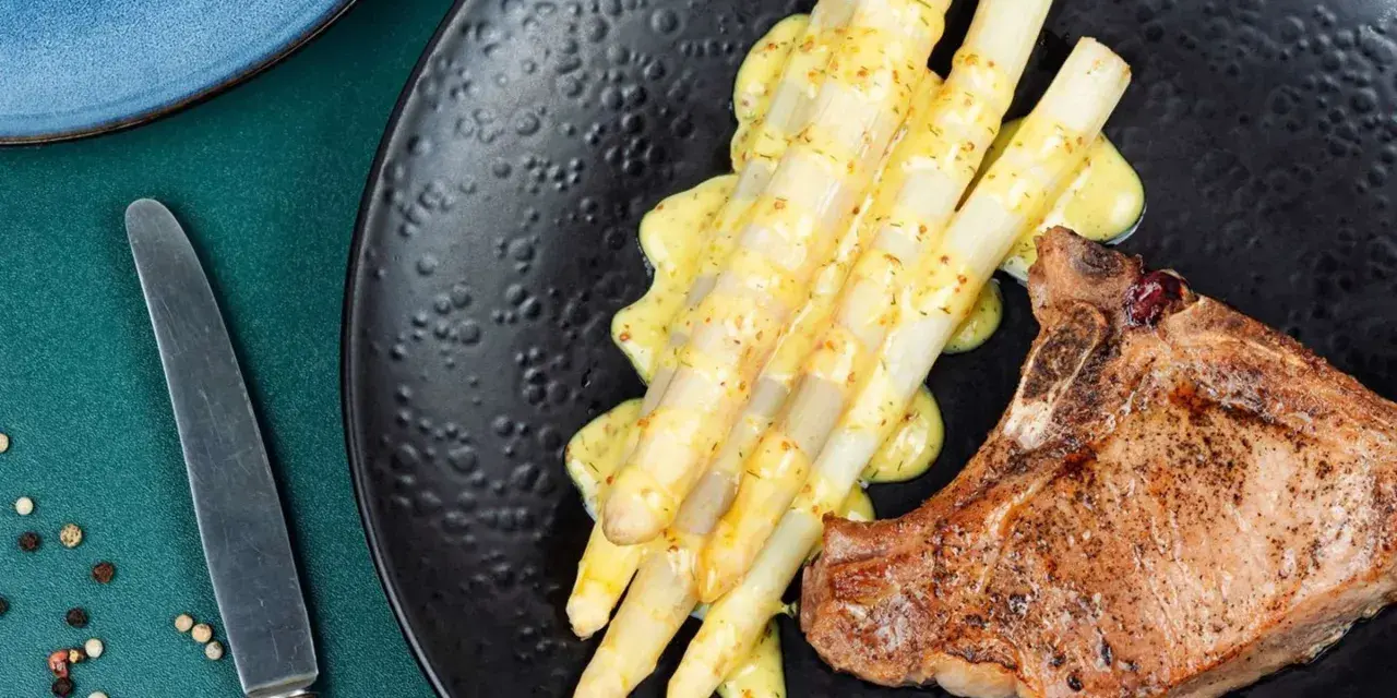 Ein saftiges Schweinekotelett liegt neben einem Haufen cremigen Spargel in der Pfanne, beträufelt mit einer würzigen Sauce.