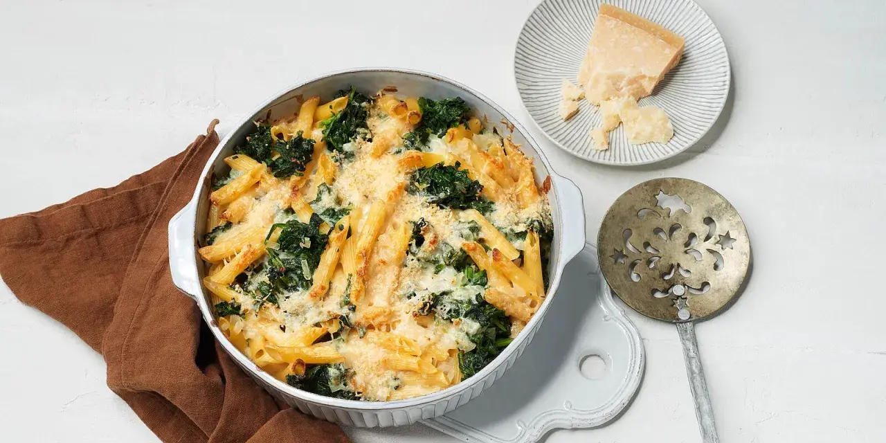Herzhafter nudelauflauf mit spinat, überbacken mit Käse. Daneben ein Stück Parmesan und ein dekoratives Servierbesteck.
