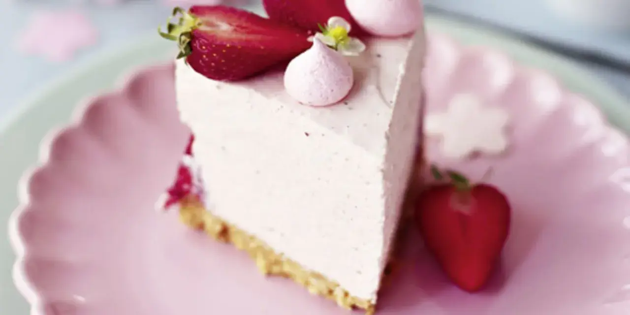 Ein Stück Erdbeer Cheesecake ohne Backen, dekoriert mit frischen Erdbeeren und rosa Baiser-Tupfern auf einem rosa Teller.