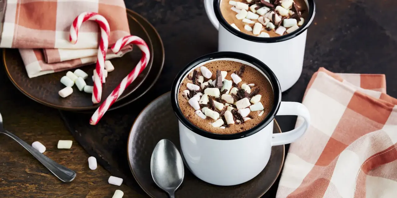 Zwei Tassen heißer Kakao mit Marshmallows und Schokostückchen, daneben Zuckerstangen und eine karierte Serviette.
