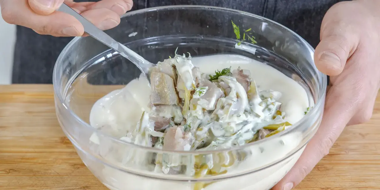 Matjes Hausfrauenart, Omas Rezept: Ein Löffel voll cremiger Sauce mit Matjesstücken und Dill wird aus einer Glasschüssel gehoben.
