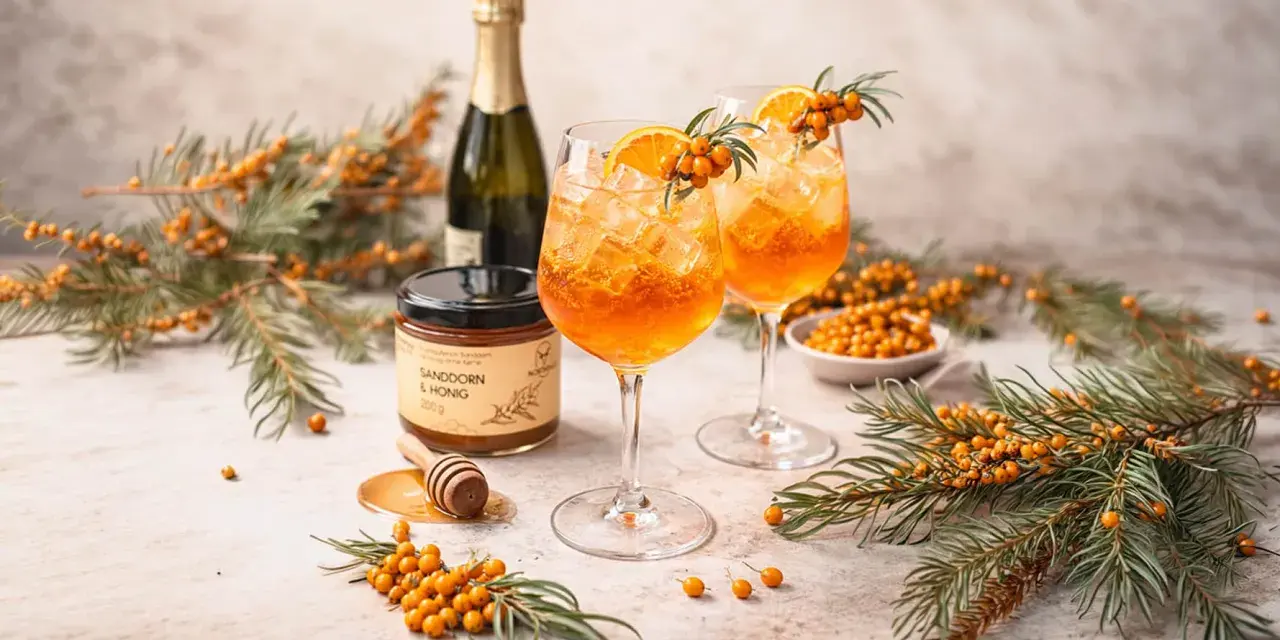 Zwei leuchtend orangefarbene Sanddorn Spritz-Cocktails mit Eis, garniert mit Orangenscheiben und Sanddornzweigen. Daneben ein Glas Sanddorn & Honig.