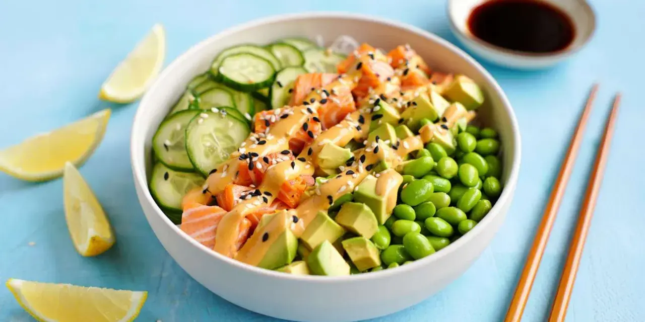 Schüssel mit einem frischen Salat aus Lachs, Avocado, Edamame und Gurkenscheiben, beträufelt mit cremiger Sauce und Sesam. Zitronenspalten und Sojasauce daneben.