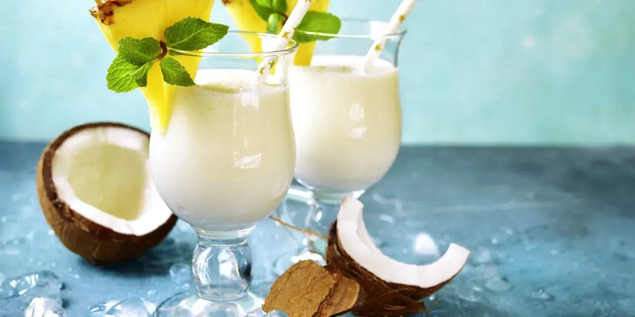 Zwei cremige Malibu Cocktails mit Ananas und Minze, umgeben von Kokosnuss und Eis.