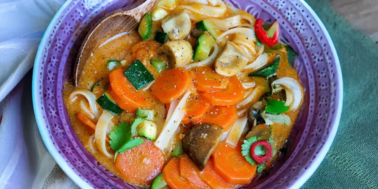 Eine dampfende, cremige Thai Curry Suppe mit Karotten, Zucchini, Pilzen und Reisnudeln in einer lila Schüssel.