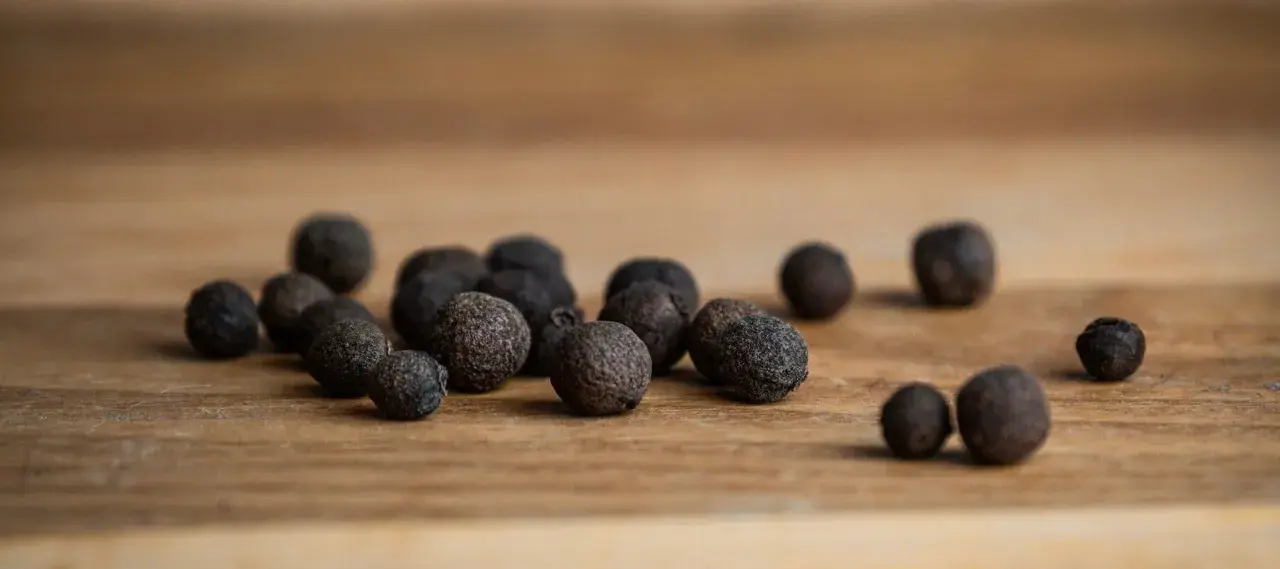 Eine Gruppe von Pimentkörnern liegt auf einem Holzbrett. Was ist Piment? Diese aromatischen Beeren duften nach Zimt, Nelken und Muskat.