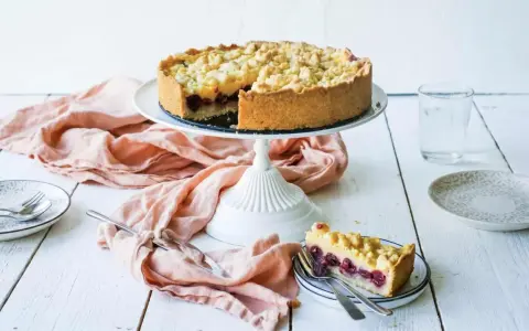 Ein saftiger Kirschkuchen mit Pudding und Streuseln auf einem Tortenständer. Ein Stück ist bereits auf einem Teller angerichtet.