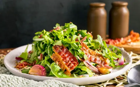 Warmer Rucolasalat mit knusprigem Speck, gerösteten Zwiebeln und Sonnenblumenkernen. Ein köstliches Gericht für Rucola Rezepte warm.