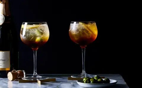 Zwei erfrischende Cynar Spritz Cocktails mit Eis, Zitrone und Oliven, daneben eine Champagnerflasche.